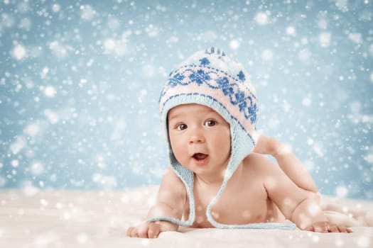 Eight month old baby in knitted winter hat lying on soft blanket with blue background and falling snow