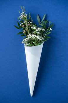 top view closeup of beautiful bouquet of delicate small white flowers with green leaves in paus tracing paper cone on blue background copyspace available