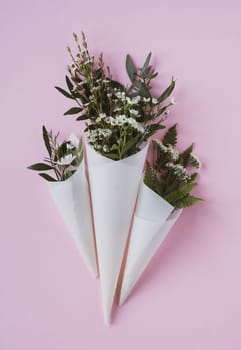 top view closeup of three beautiful bouquets of delicate small white flowers with green leaves in paus tracing paper cone on pink background copyspace available