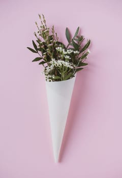 top view closeup of beautiful delicate small white flowers with green leaves in paus tracing paper cone on pink background copyspace available