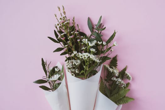 top view closeup of three beautiful bouquets of delicate small white flowers with green leaves in paus tracing paper cone on pink background copyspace available