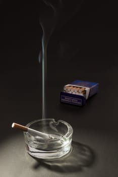 A lid cigarette lies burning in an ashtray, with the smoke floating up in to the air, with an open packet of cigarettes in the background.