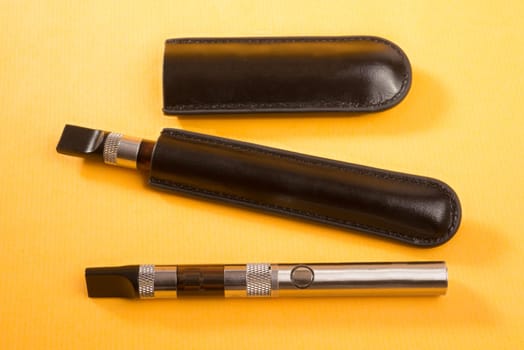 Two electronic cigarettes lie on a yellow background, with one unit still in leather pouch.
