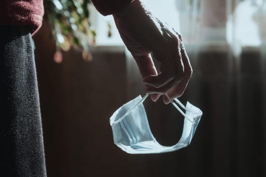Old woman holding face mask in hand standing near window in her home.