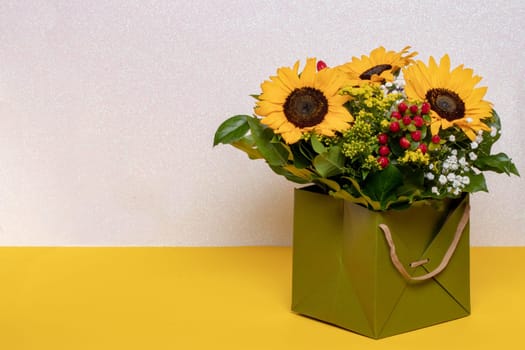 Closeup of a beautiful bouquet with yellow sunflowers in a decorative green gift box on a yellow table over a light pink glittering background. Macro. Space for design. Card concept.