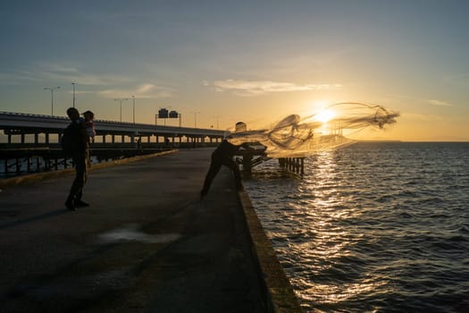 Batu Maung, Penang/Malaysia - Jan 01 2020: A man casting net to sea. Another man hug a child.