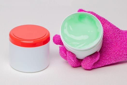Close-up of a closed and an open container with cooling gel (horse balm) for sports injuries on a light background. The open container is held by a pink massage glove. Copy space. Macro.