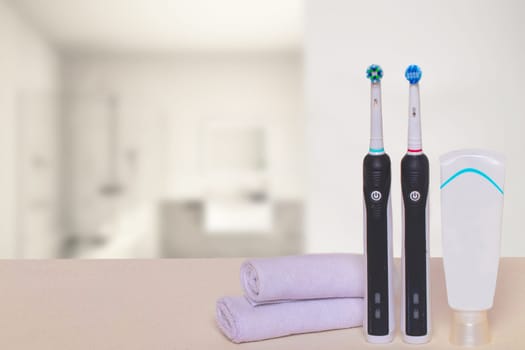 Two electrical tooth brushes, an oral care product or toothpaste and towels on table against blurred bright bathroom background. Dental and healthcare concept. Copy space.
