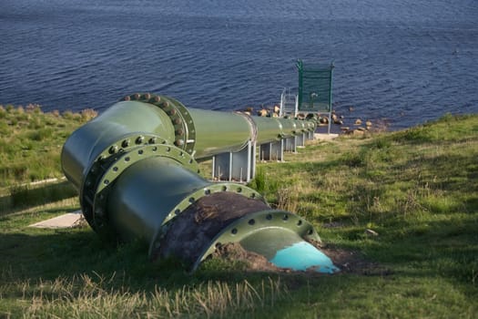 Full frame image of large green metal pipe in an outdoor hydro dam setting. Modern and bolted together, with control equipment at the reservoir end