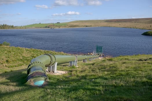 Warm sunlight shines on a shiny green pipe feeding water into a deep blue rural reservoir. Cloudy sky background. Modern industrial hydro equipment.