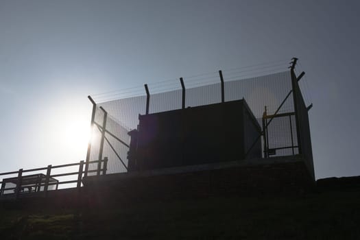 Tall barbed wire anti climb fence encases large square metal industrial water management equipment. Looking up, plain blue sky and bright sun behind