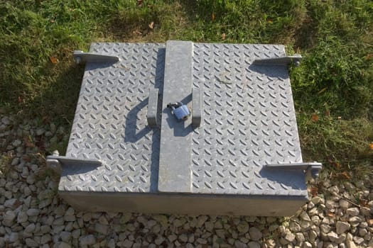 Modern secure padlocked steel service hatch in rural sunlight. Manhole cover juxtaposed against natural surroundings. Shadowed checkered steel texture