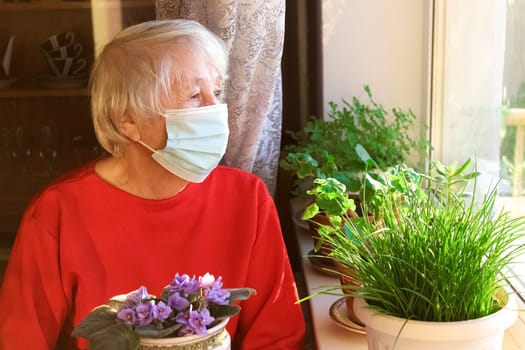 The Covid-19, health, safety and pandemic concept - senior old lonely woman wearing protective medical mask sitting near the window at home for protection from virus