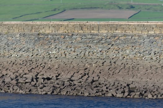 A rural large stone dam acts as the wall to a deep blue water reservoir. Selective focus blurs the agricultural fields in the background. Warm sun.