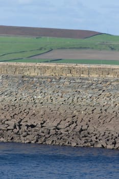 Vertical image of stone dam reservoir wall. Deep blue water and rural agricultural background are selectively out of focus. Warm sunny summers day