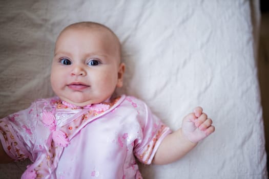 Curious adorable baby girl from above in Asian pink attire lying down on white bed. Portrait of cute female baby resting on bed. Happy babies on cradles and beds