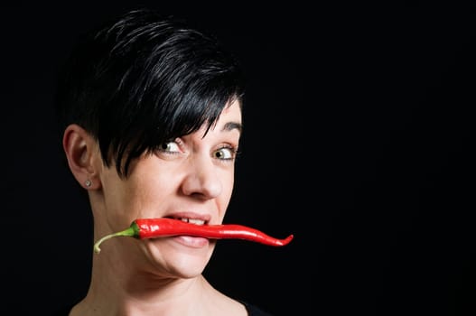 Lateral head portrait of a young black-haired woman in front of a black background holding a long red chilli pod across her teeth and looking skeptically into the camera.