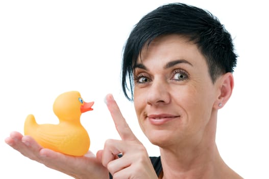 Head-and-shoulder view of a young black-haired woman holding a rubber duck on her right hand at chin level and holding a white cream swab with the index finger of the left hand in the direction of the rubber duck and looking friendly into the camera against white background.