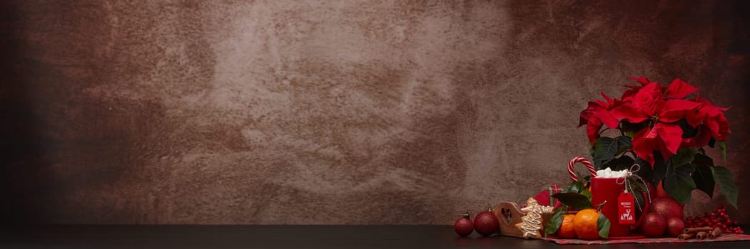 Red Poinsettia flowers, tray with ginger cookies and mandarines, mug with hot cocoa and marshmallows. fir ornaments on brown background with copy space. Winter holidays. New Year, Christmas treat.