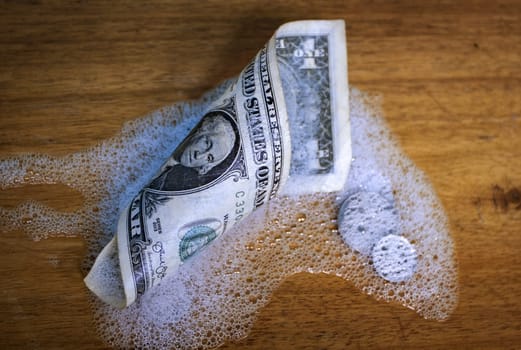 A rolled up dollar bill and coins, partially covered by foam on a wooden surface