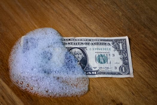 A one dollar bill partially covered by foam on a wooden surface