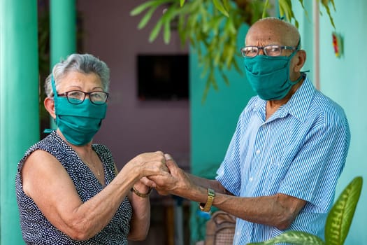 An elderly couple, both wearing face mask and glasses and holding hands