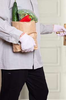 A man wearing medical gloves and a shopping bag with fresh vegetables enters the house. Concept of strengthening the immune system and protection against coronavirus infections.