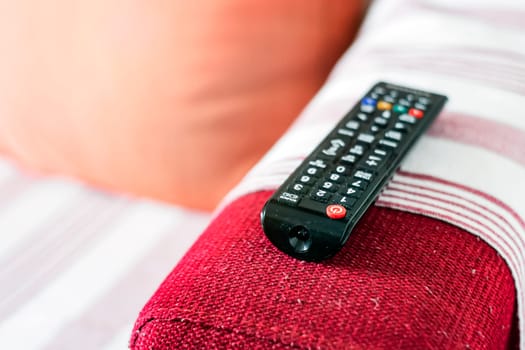 the dusty television remote control on the armrest of a red fabric sofa. Lint on the sofa. Sedentary life and home entertainment. Selective focus on the remote control