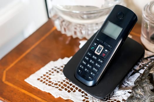 a black plastic cordless phone charging on its cradle inside a classically furnished home with antiques and white crochet. Outdated technology