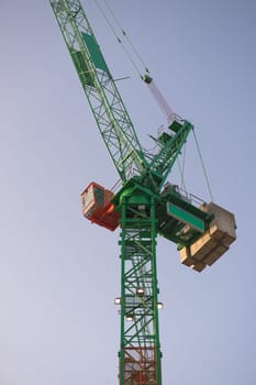 Vertical sunrise image looking up at a tall green and orange tower crane. Full frame crane and cabin with a plain blue sky background.