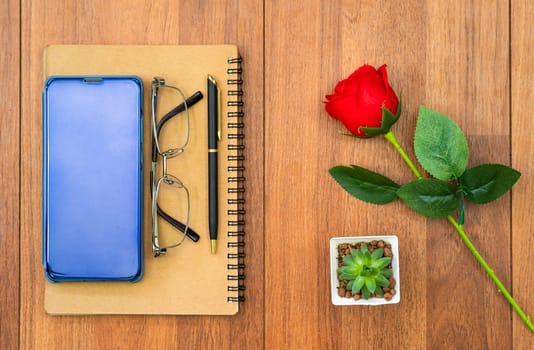 Top view image of notebook and cellphone with red rose on wooden table background