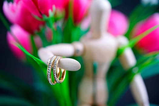 Wooden figure holding two golden wedding ring resting with fake pink tulip flower