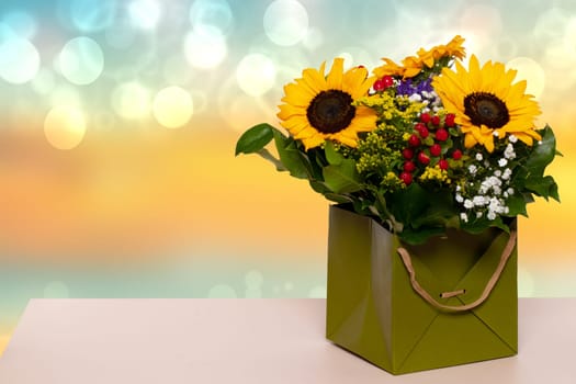 Closeup of a beautiful bouquet with yellow sunflowers in a decorative green gift box on a yellow table over a blurred blue yellow background. Macro. Space for design. Card concept.