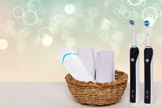Two electrical tooth brushes, an oral care product or toothpaste and towels in a basket on a table against abstract blurred bright water bubbles background. Dental and healthcare concept. Copy space.
