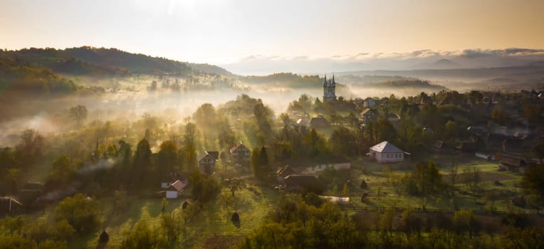 Aerial view of village in fog with golden sunbeams at sunrise in autumn. Beautiful rural landscape with foggy colorful trees, church, orange sky with sun. Fall in Romania.