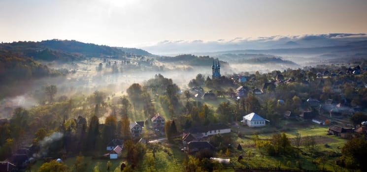 Aerial view of village in fog with golden sunbeams at sunrise in autumn. Beautiful rural landscape with foggy colorful trees, church, orange sky with sun. Fall in Romania.