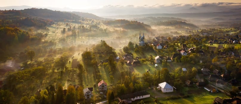 Aerial view of village in fog with golden sunbeams at sunrise in autumn. Beautiful rural landscape with foggy colorful trees, church, orange sky with sun. Fall in Romania.