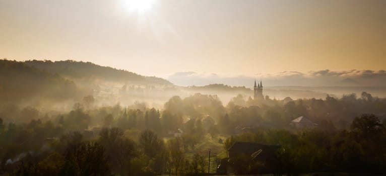 Aerial view of village in fog with golden sunbeams at sunrise in autumn. Beautiful rural landscape with foggy colorful trees, church, orange sky with sun. Fall in Romania.