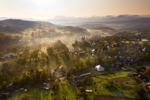 Aerial view of village in fog with golden sunbeams at sunrise in autumn. Beautiful rural landscape with foggy colorful trees, church, orange sky with sun. Fall in Romania.