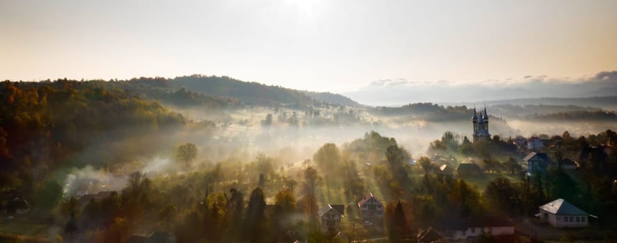 Aerial view of village in fog with golden sunbeams at sunrise in autumn. Beautiful rural landscape with foggy colorful trees, church, orange sky with sun. Fall in Romania.
