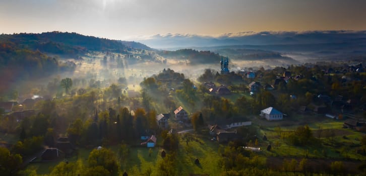 Aerial view of village in fog with golden sunbeams at sunrise in autumn. Beautiful rural landscape with foggy colorful trees, church, orange sky with sun. Fall in Romania.