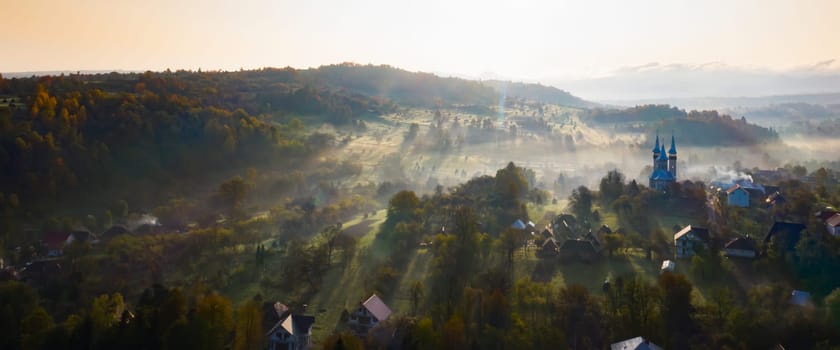 Aerial view of village in fog with golden sunbeams at sunrise in autumn. Beautiful rural landscape with foggy colorful trees, church, orange sky with sun. Fall in Romania.