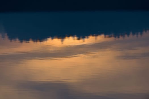 A warm orange glowing sunset behind a line of conifer trees is reflected and displayed upside down in the calm ripples of Redmires reservoir.
