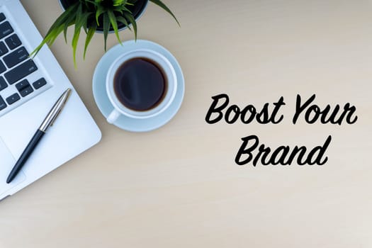 BOOST YOUR BRAND text with fountain pen, laptop, decorative flower and cup of coffee on wooden background. Business and copy space concept..