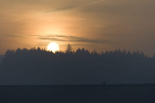 Three people and a dog walk along the top of a dam wall at redmires reservoirs near sheffield. Behind them a hazy golden sunset silhouettes a forest