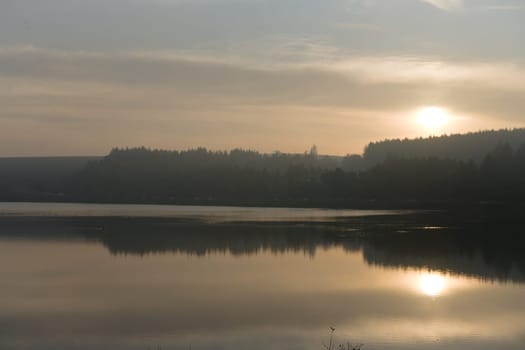 Over the calm evening water of redmires reservoirs, a long line of cars are parked in front of a large forest, underneath the warm sunset glow.