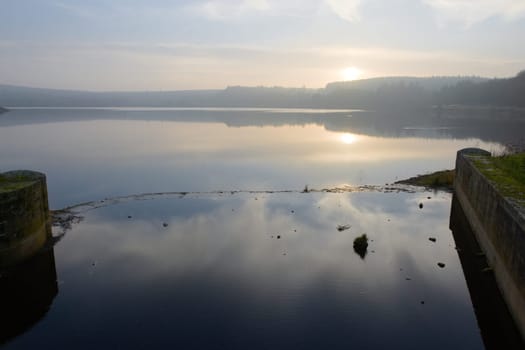 Calm background horizontal landscape image of a sunset reflected in a reservoir near sheffield. Brick walls of reservoir inlet in the foreground.