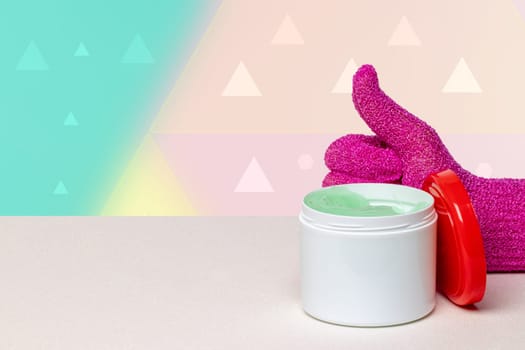 Container with green cooling gel for sports injuries and a pink massage glove with a thumbs up on bright table against abstract pastel background. Space.