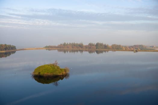 Small white birds rest on the still waters surface at redmires reservoirs. Dam wall circles the water, and an unusual island is the foreground.