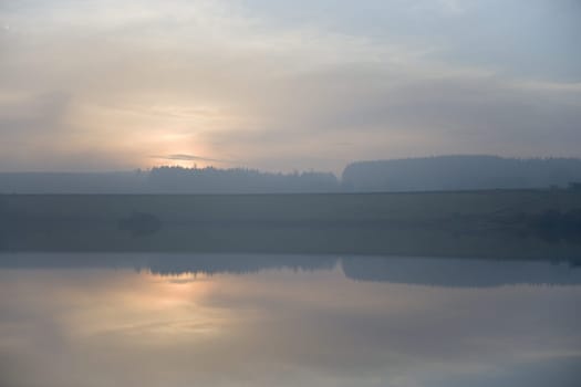 Calm reservoir water reflects misty sunset. Serene horizontal landscape image with blue and orange pastel colors. Hazy atmosphere with wispy clouds